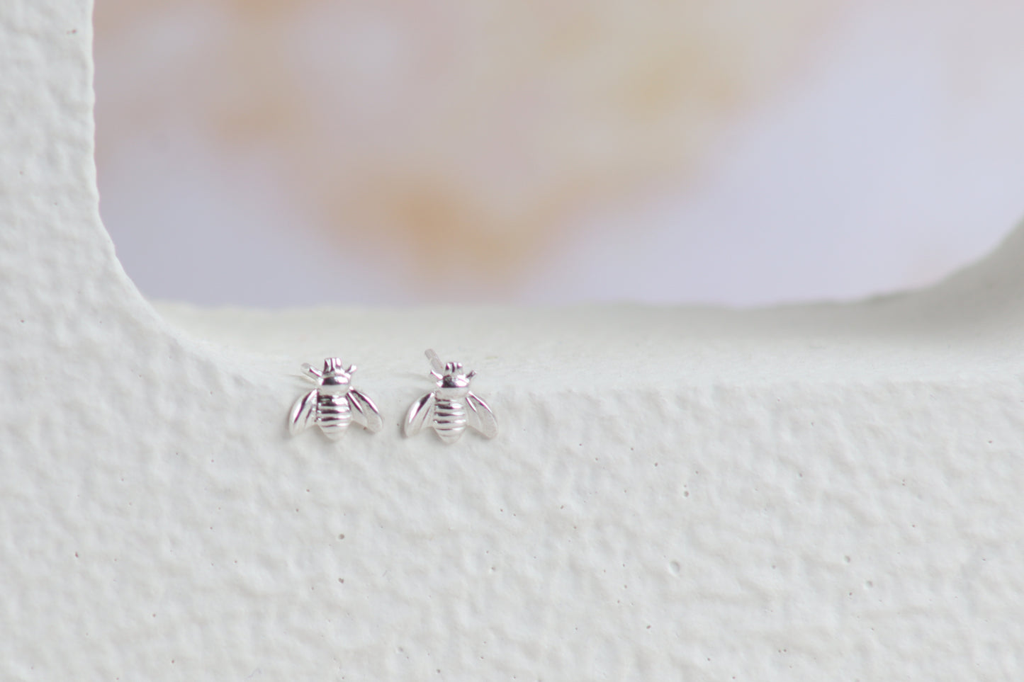 A pair of tiny silver bee stud earrings resting on a textured white surface with a soft, blurred light background.