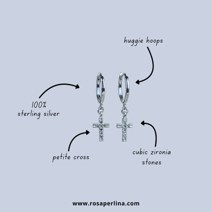 Silver hoop earrings with crosses and cubic zirconia stones on a light gray background, labeled as 'huggie hoops' and '100% sterling silver'.