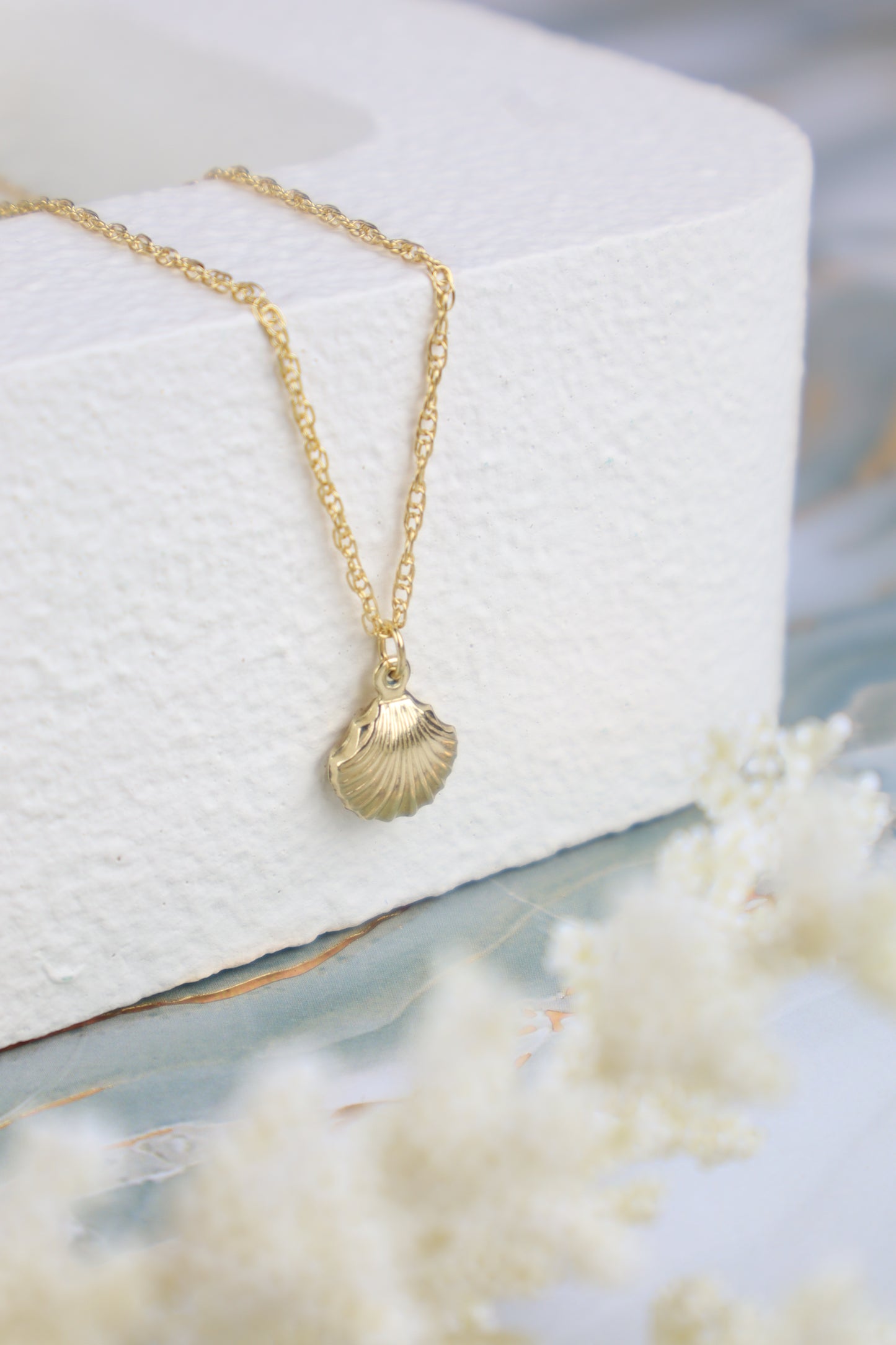 Gold necklace with a shell pendant on a white cushion, surrounded by white flowers.