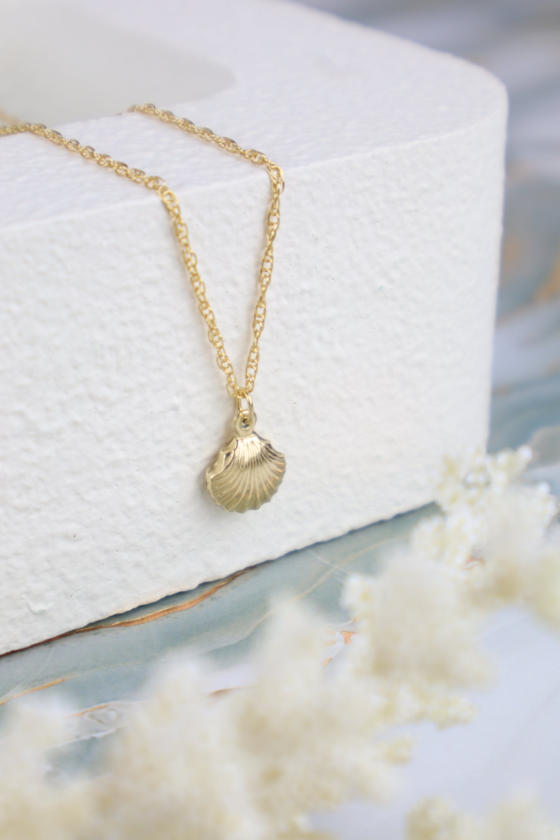 Gold necklace with a shell pendant on a white cushion, surrounded by white flowers.