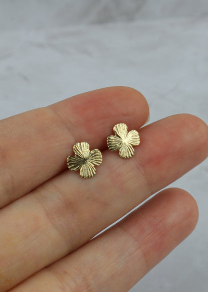A pair of textured gold four-leaf clover stud earrings held on a person's fingers against a light gray marble background.