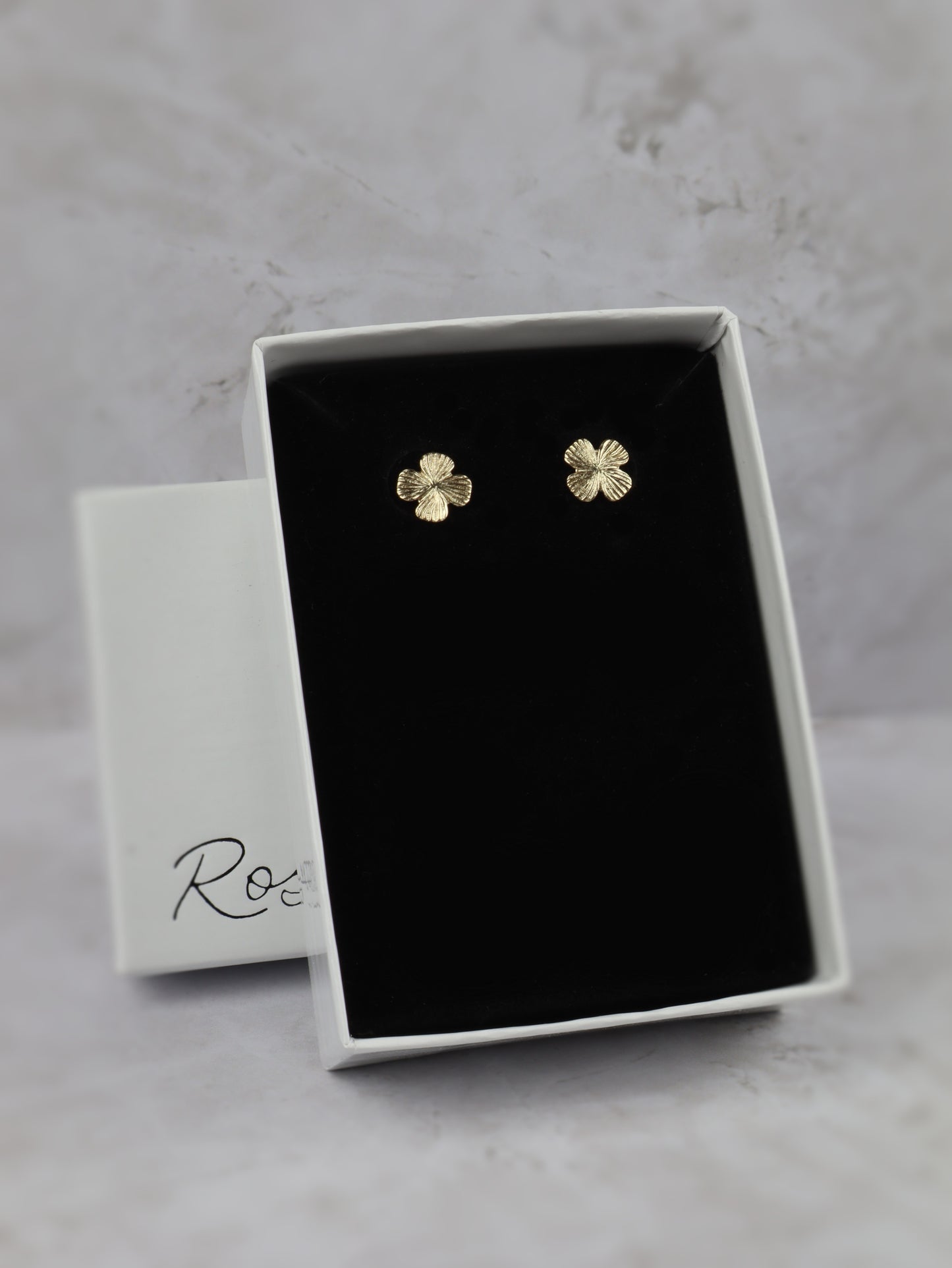 A pair of small gold four-leaf clover stud earrings displayed in an open white jewelry box with a black velvet insert, set against a light gray marble-like background. 