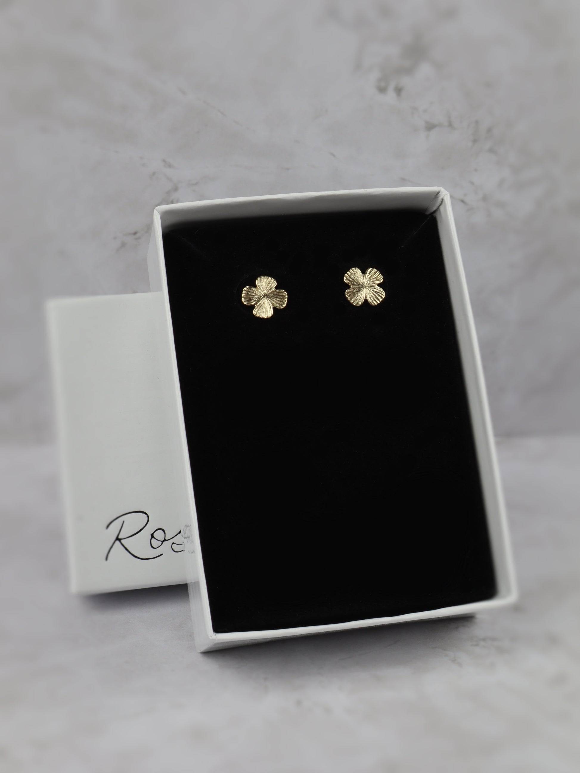 A pair of small gold four-leaf clover stud earrings displayed in an open white jewelry box with a black velvet insert, set against a light gray marble-like background. 