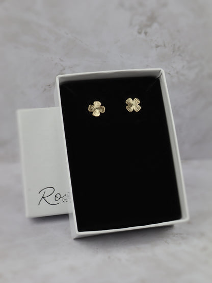 A pair of small gold four-leaf clover stud earrings displayed in an open white jewelry box with a black velvet insert, set against a light gray marble-like background. 