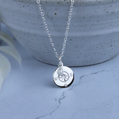 A silver necklace with a round compass pendant and a small pearl accent, displayed on a light blue marble surface in front of a speckled ceramic pot.