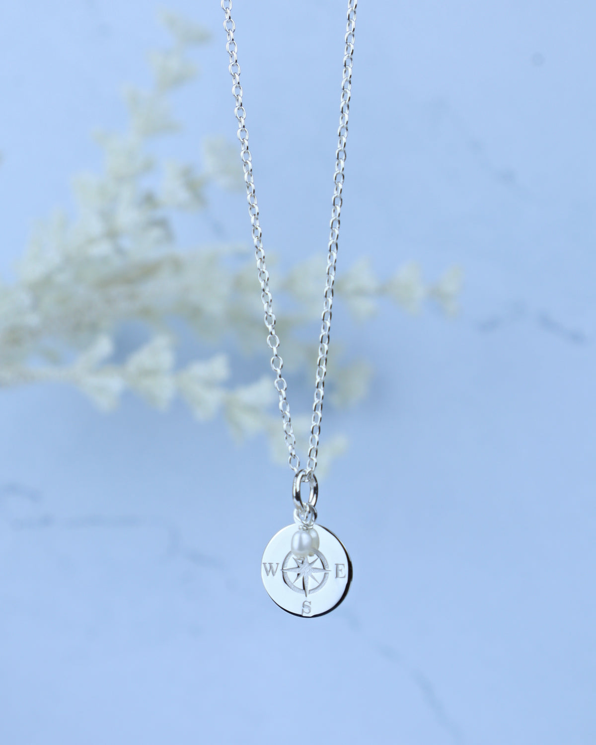 A silver chain necklace with a round silver disc pendant engraved with a compass rose and a small pearl at the center, shown against a soft light blue background with a blurred white floral element.