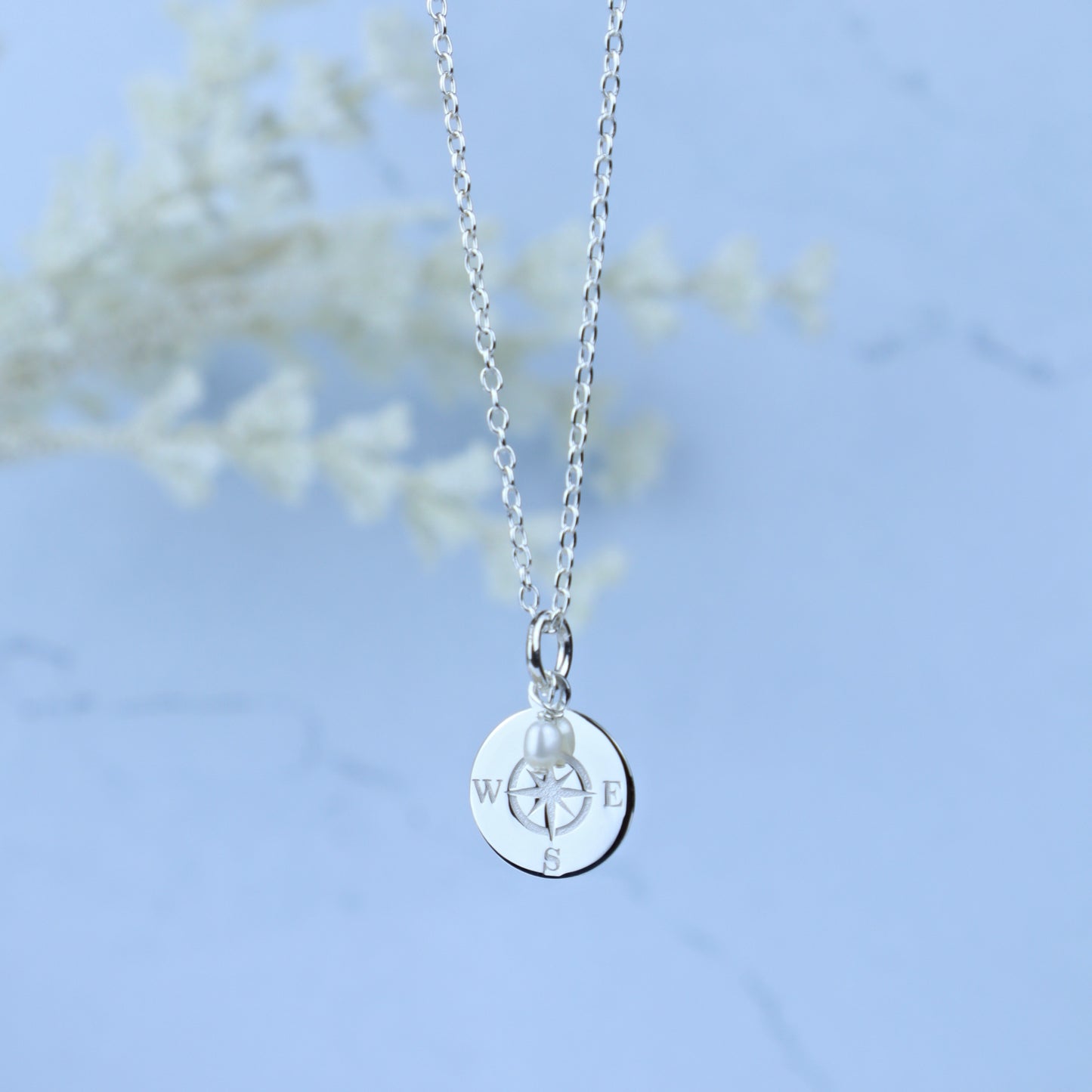 A silver chain necklace with a round silver pendant engraved with compass directions and a small pearl bead, shown against a soft light blue background with blurred white floral decor.