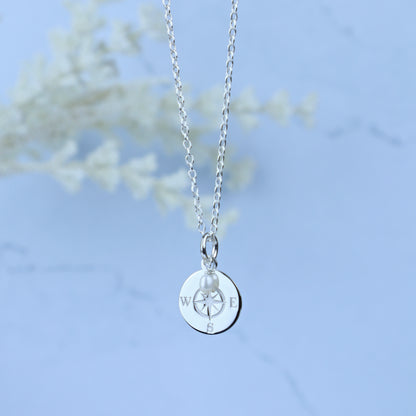 A silver chain necklace with a round silver pendant engraved with compass directions and a small pearl bead, shown against a soft light blue background with blurred white floral decor.