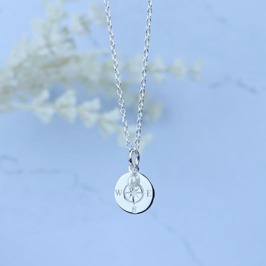 A silver chain necklace with a round silver pendant engraved with compass directions and a small pearl bead, shown against a soft light blue background with blurred white floral decor.
