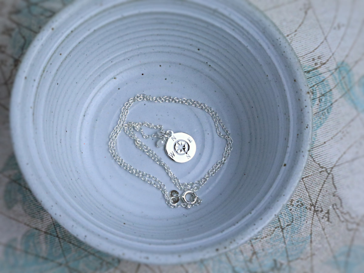 A silver chain necklace with a round engraved disc pendant and a small key-shaped charm, displayed inside a light blue speckled ceramic bowl.