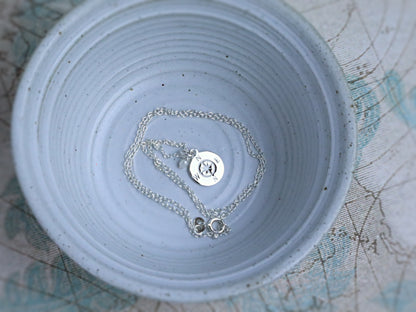 A silver chain necklace with a round engraved disc pendant and a small key-shaped charm, displayed inside a light blue speckled ceramic bowl.