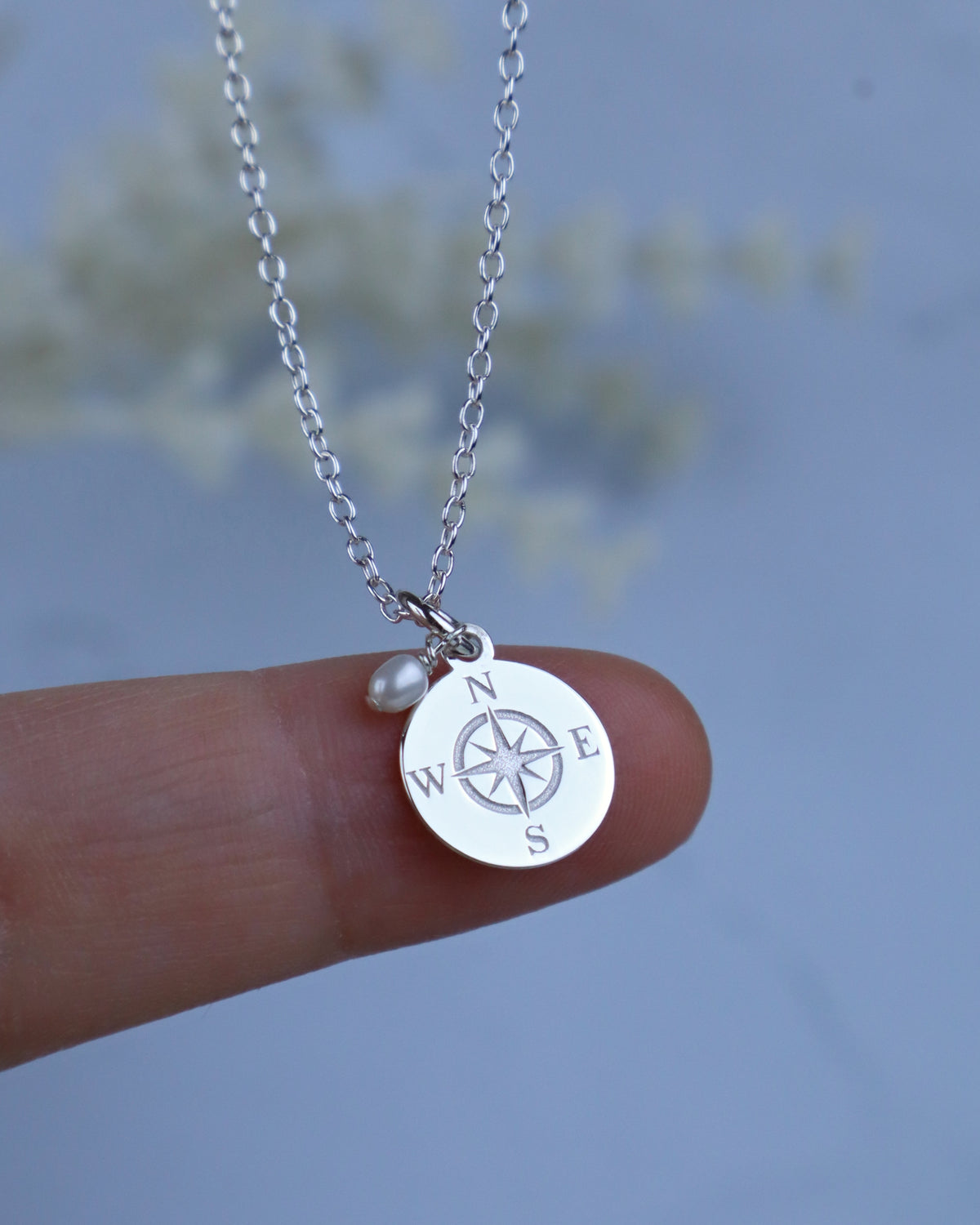 A silver compass pendant necklace held on a person's finger, featuring a round compass disc and a small pearl bead on a delicate chain against a soft blue background.
