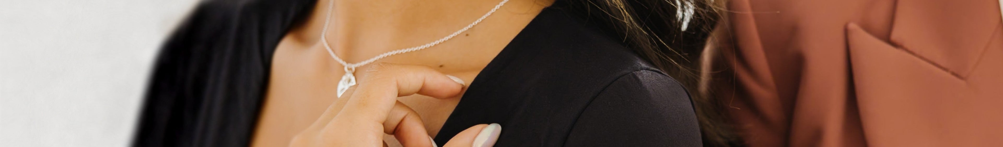 A close-up lifestyle shot of a model wearing a delicate silver necklace with a small clear heart-shaped pendant, paired with a black top.