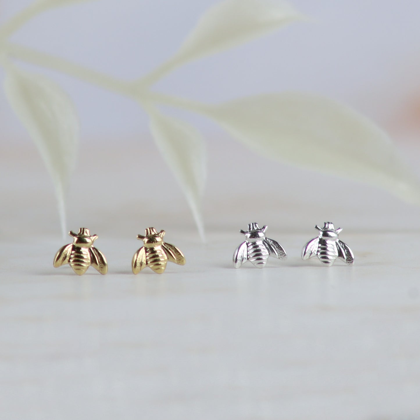 Four small bee-shaped stud earrings displayed on a light surface, with two gold-tone bees on the left and two silver-tone bees on the right, against a softly blurred pale background with faint leaf shapes.