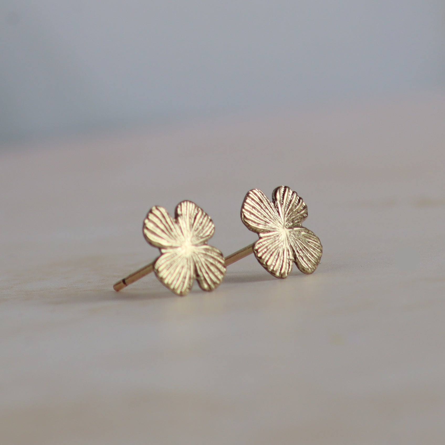 A pair of small gold four-leaf clover stud earrings with textured petals, resting on a light beige surface with a soft-focus gray background.