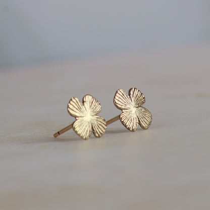 A pair of small gold four-leaf clover stud earrings with textured petals, resting on a light beige surface with a soft-focus gray background.