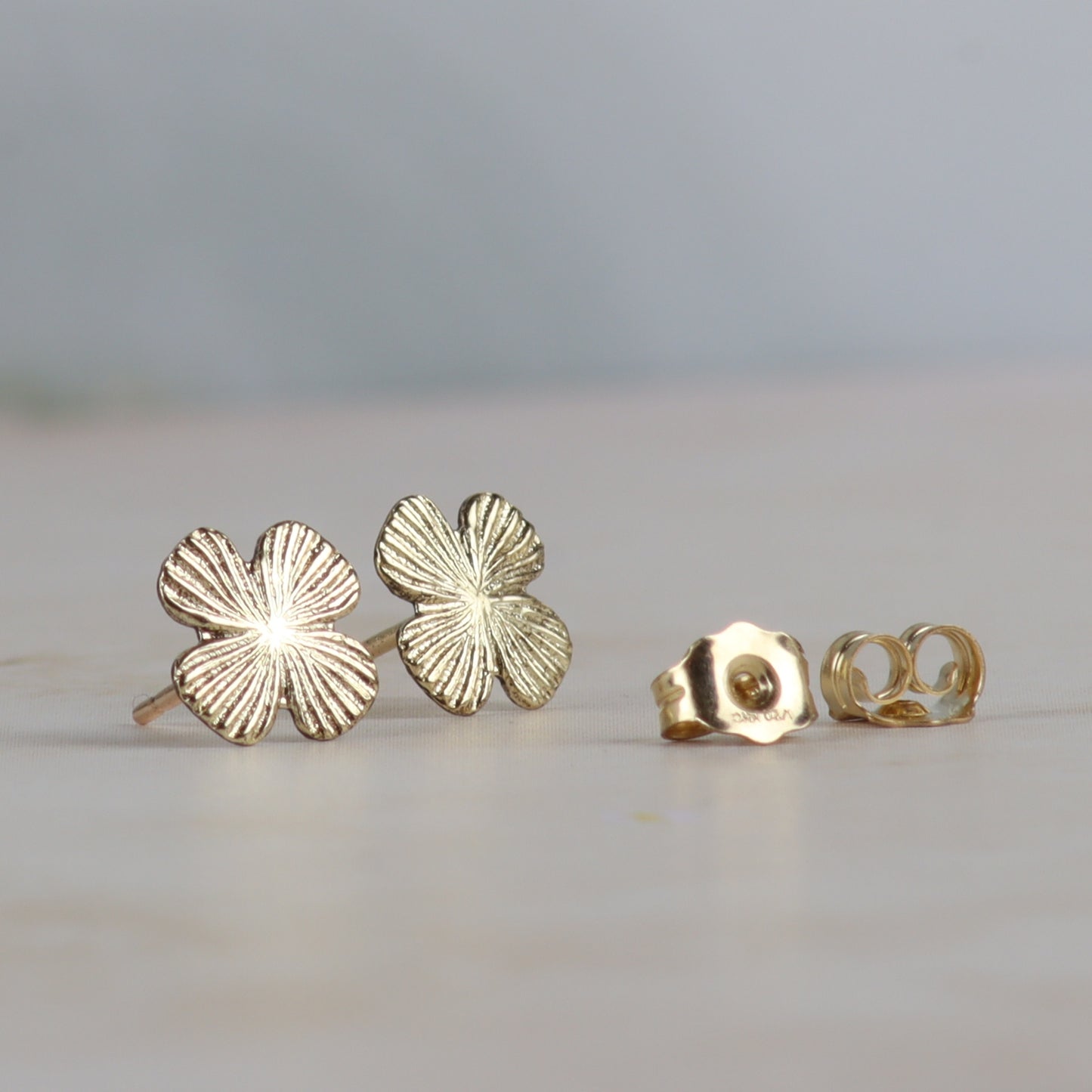 A pair of gold four-leaf clover stud earrings with textured petals, shown next to their gold flat-back post backs on a light beige surface with a soft gray background.