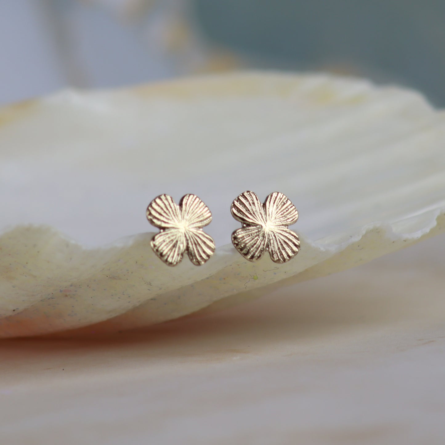 A pair of small four-petal flower stud earrings in a textured rose gold finish, resting on a smooth, light-colored shell against a soft-focus background.