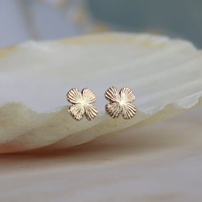 A pair of small four-petal flower stud earrings in a textured rose gold finish, resting on a smooth, light-colored shell against a soft-focus background.