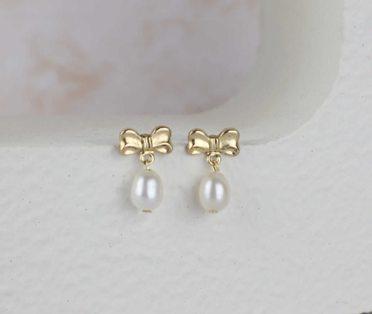 A pair of gold bow stud earrings with dangling white freshwater pearls, displayed on a textured white surface.