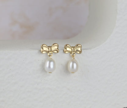 A pair of gold bow stud earrings with dangling white freshwater pearls, displayed on a textured white surface.