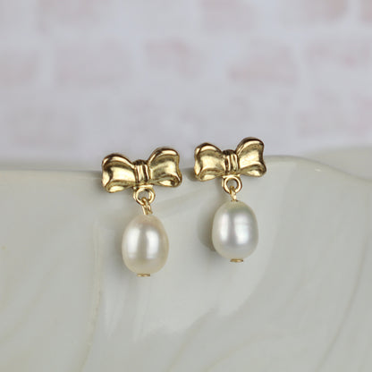 A pair of gold bow stud earrings with dangling white pearl drops, displayed on a light-colored marble surface.