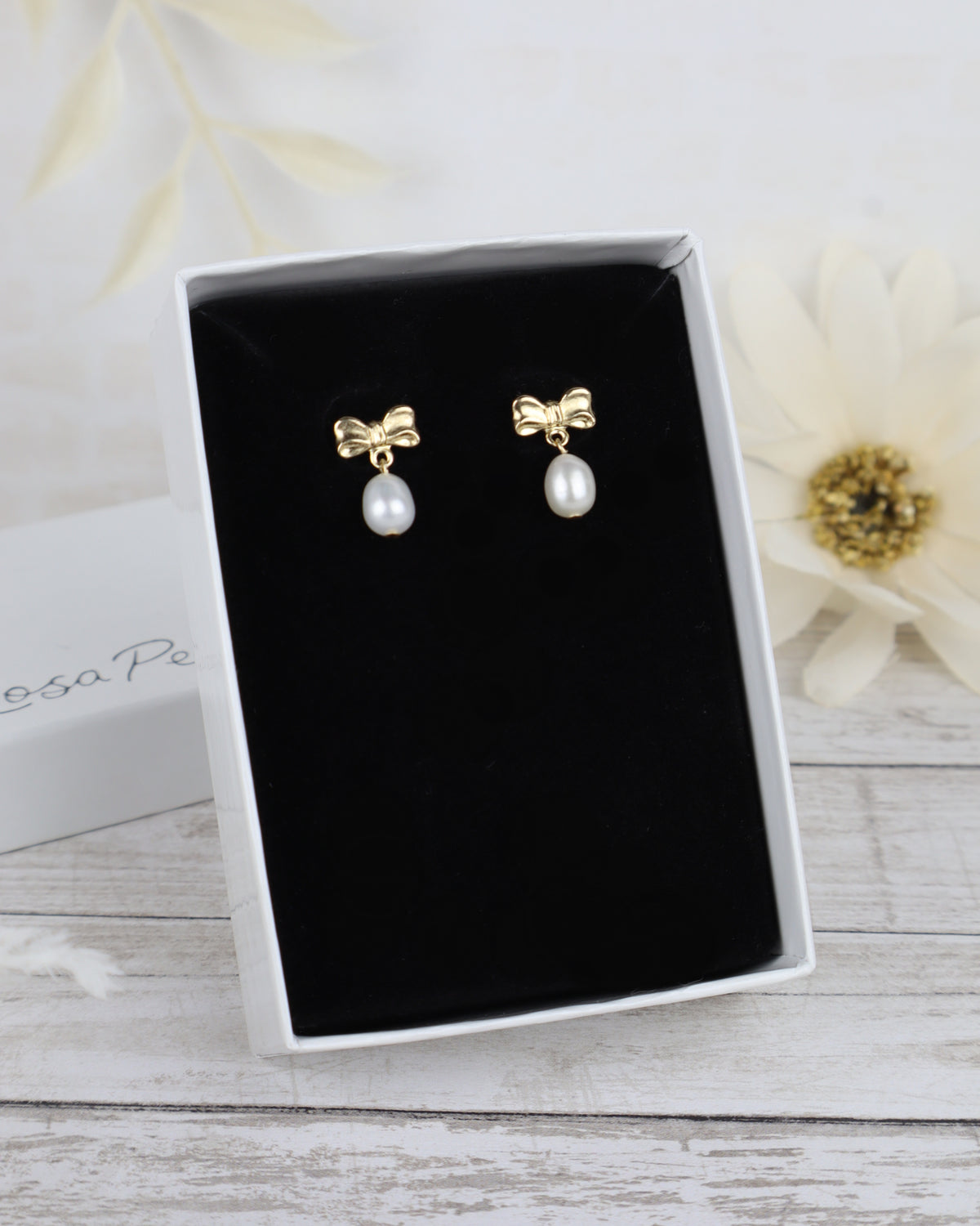 A pair of gold bow stud earrings with dangling white pearl drops, presented in an open black jewelry box on a light wood surface with a blurred white flower in the background.