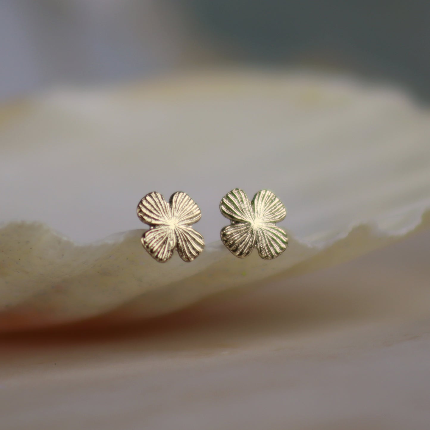 A pair of small gold four-petal flower stud earrings resting on a smooth, light beige stone surface with a soft-focus background.