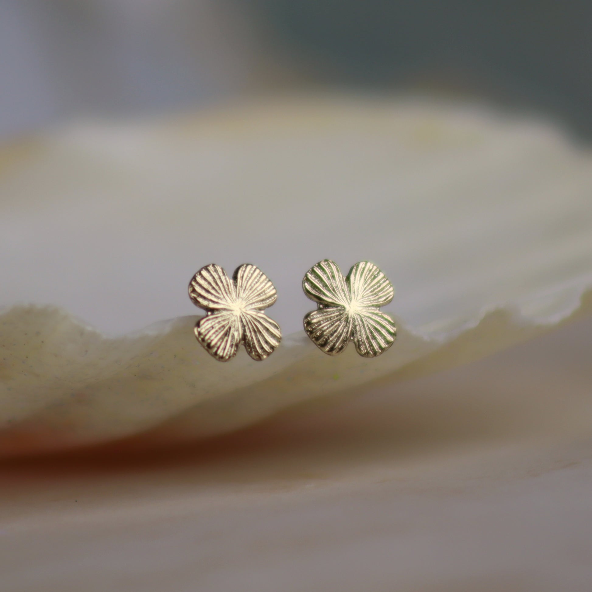 A pair of small gold four-petal flower stud earrings resting on a smooth, light beige stone surface with a soft-focus background.