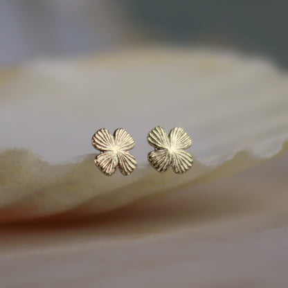 A pair of small gold four-petal flower stud earrings resting on a smooth, light beige stone surface with a soft-focus background.