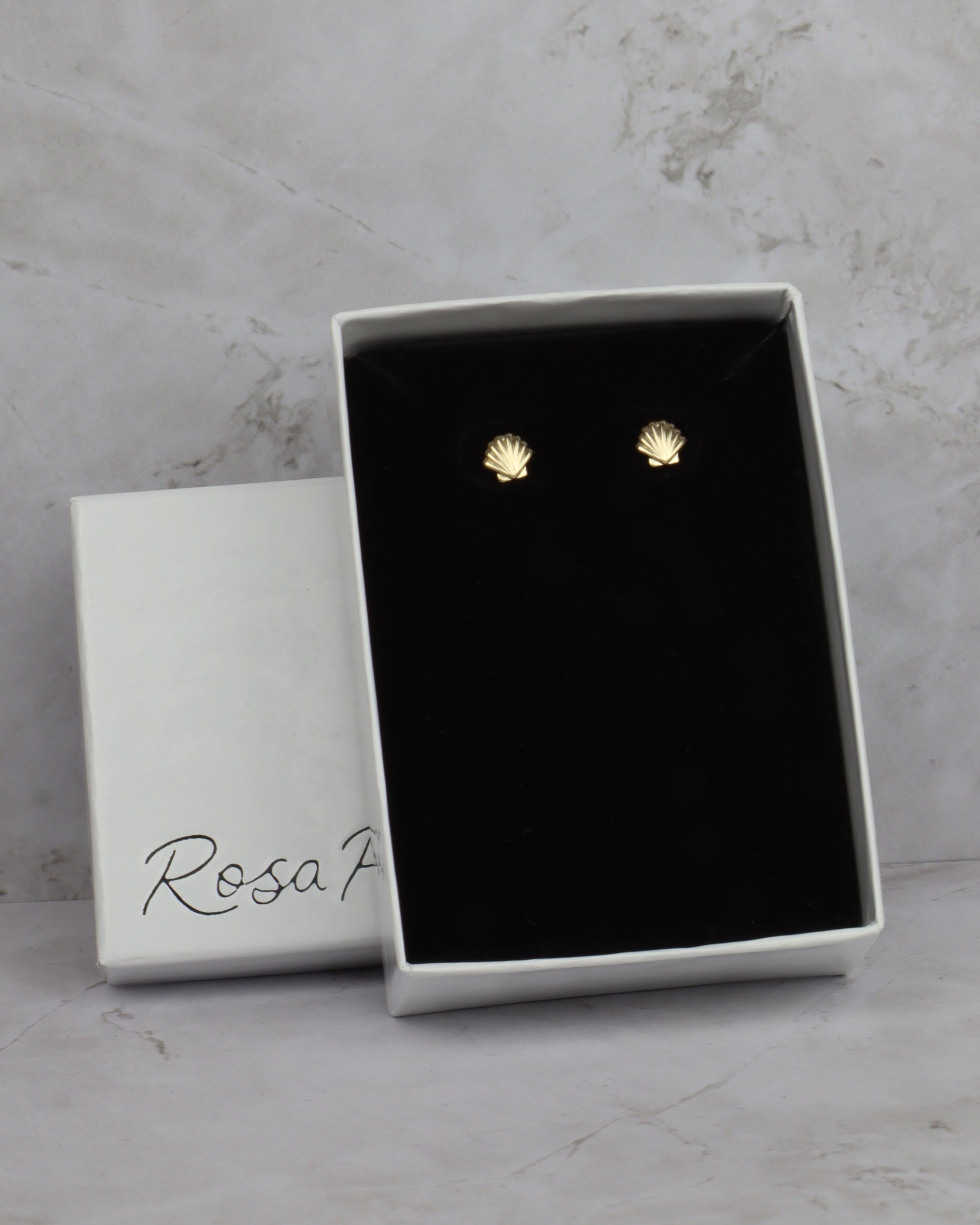 A pair of small gold stud earrings shaped like seashells, presented in an open white jewelry box with a black velvet insert. The closed white box lid beside it has the brand name 'Rosa F' printed on it, set against a light gray marble background.