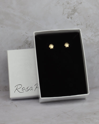 A pair of small gold stud earrings shaped like seashells, presented in an open white jewelry box with a black velvet insert. The closed white box lid beside it has the brand name 'Rosa F' printed on it, set against a light gray marble background.