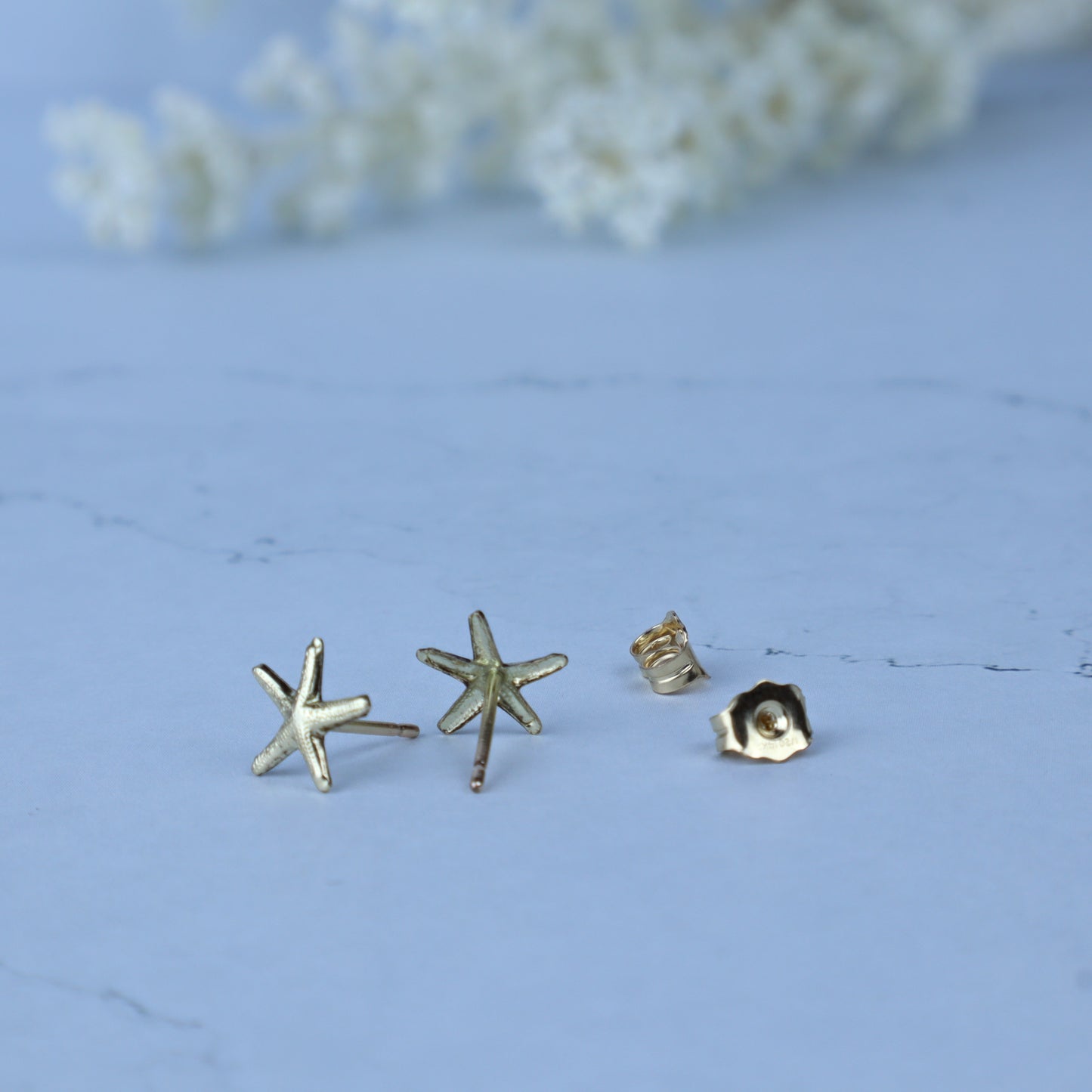 Three small gold-toned stud earrings on a pale blue surface, including two starfish-shaped earrings and one flower-shaped earring, with a soft blurred floral background.