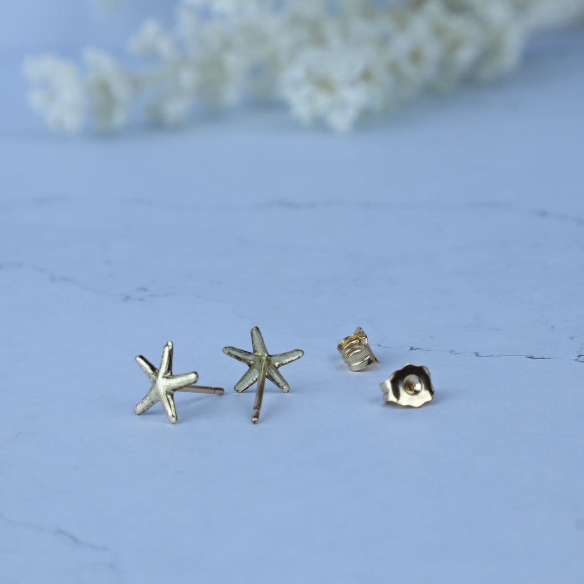 Three small gold-toned stud earrings on a pale blue surface, including two starfish-shaped earrings and one flower-shaped earring, with a soft blurred floral background.