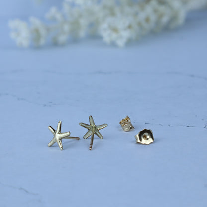 Three small gold-toned stud earrings on a pale blue surface, including two starfish-shaped earrings and one flower-shaped earring, with a soft blurred floral background.
