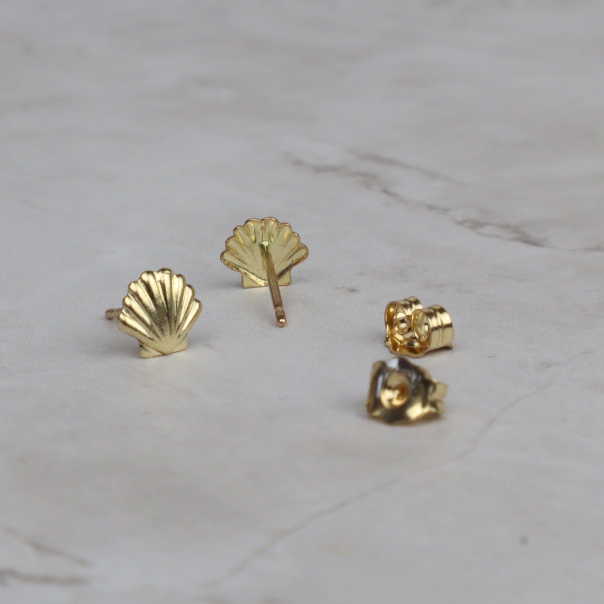 Four small gold stud earrings arranged on a light marble surface, including two shell-shaped studs, one rose-shaped stud, and one close-up of the metal backing.