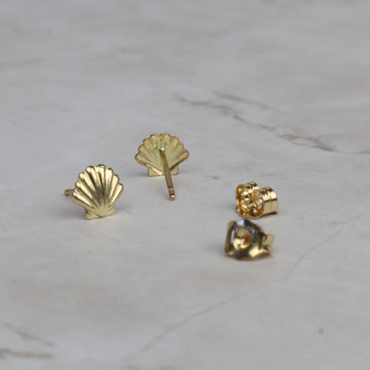 Four small gold stud earrings arranged on a light marble surface, including two shell-shaped studs, one rose-shaped stud, and one close-up of the metal backing.