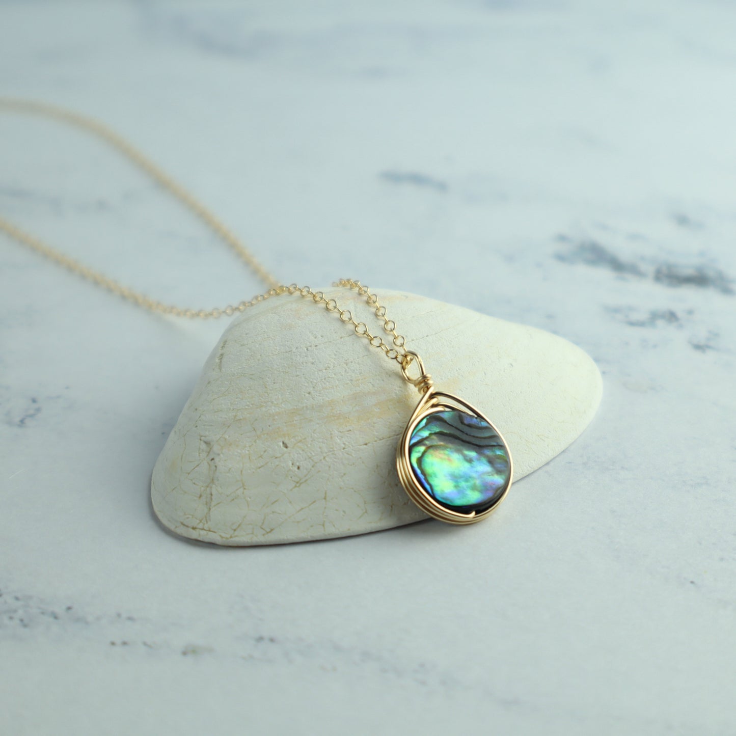 Gold necklace with a teardrop-shaped abalone shell pendant on a marble surface