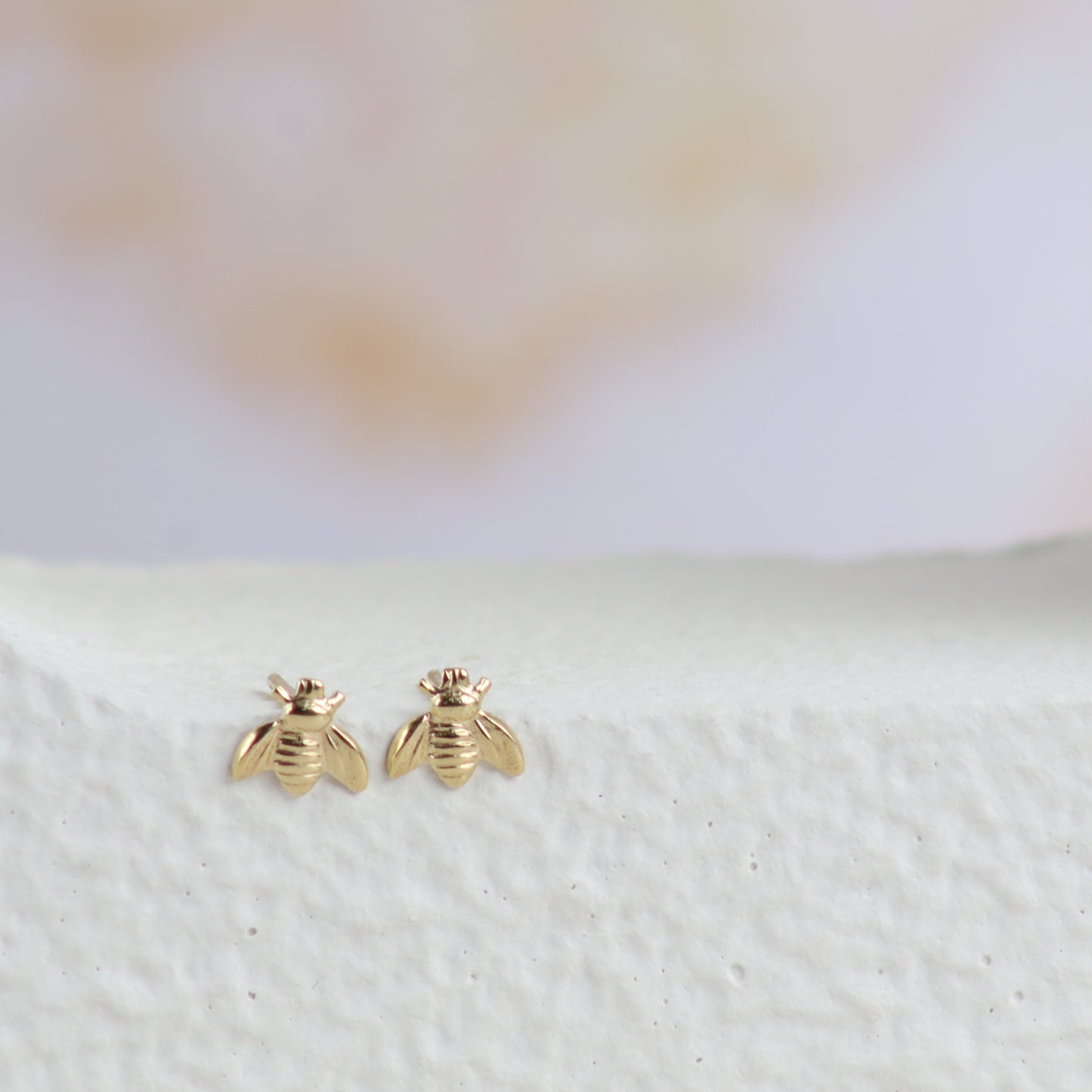 A pair of small gold bee stud earrings displayed on a textured white surface with a soft, blurred light background.