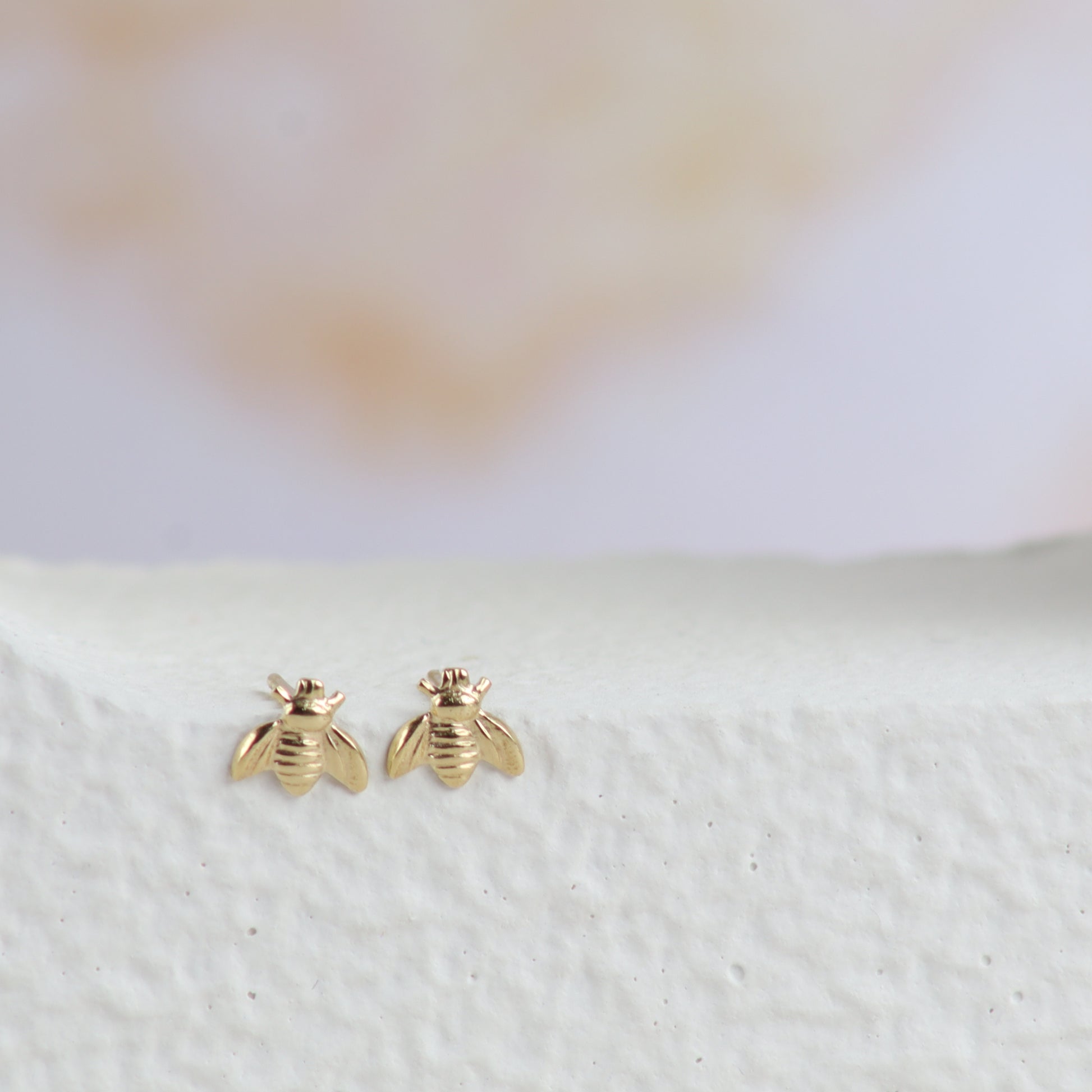A pair of small gold bee stud earrings displayed on a textured white surface with a soft, blurred light background.