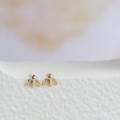 A pair of small gold bee stud earrings displayed on a textured white surface with a soft, blurred light background.