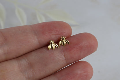 A close-up of a person's hand holding a pair of small gold bee stud earrings against a soft light gray background.