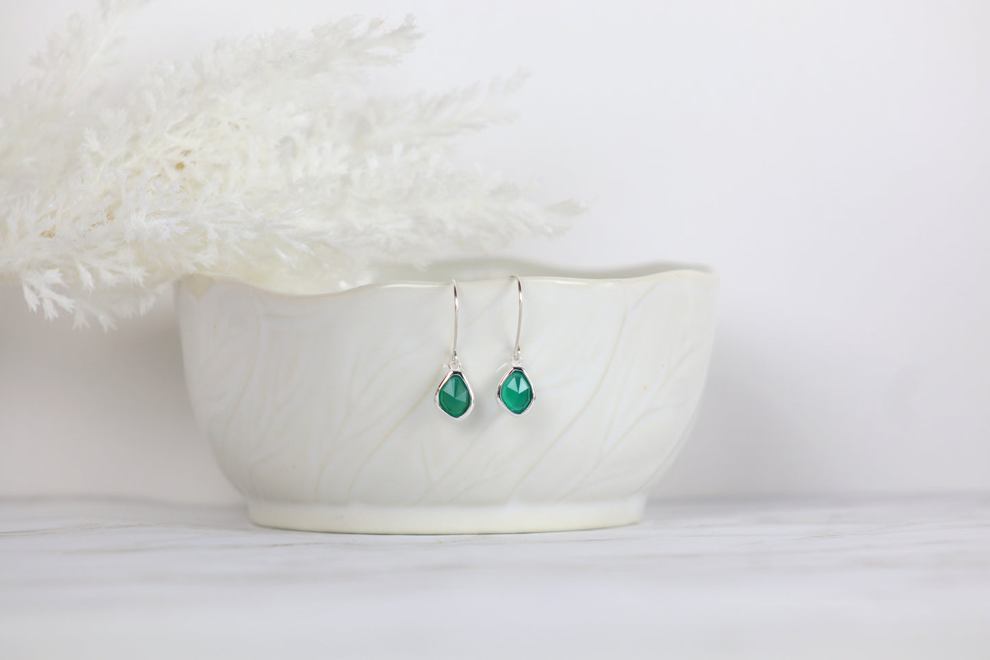 Green teardrop earrings hanging from a white bowl with a white background