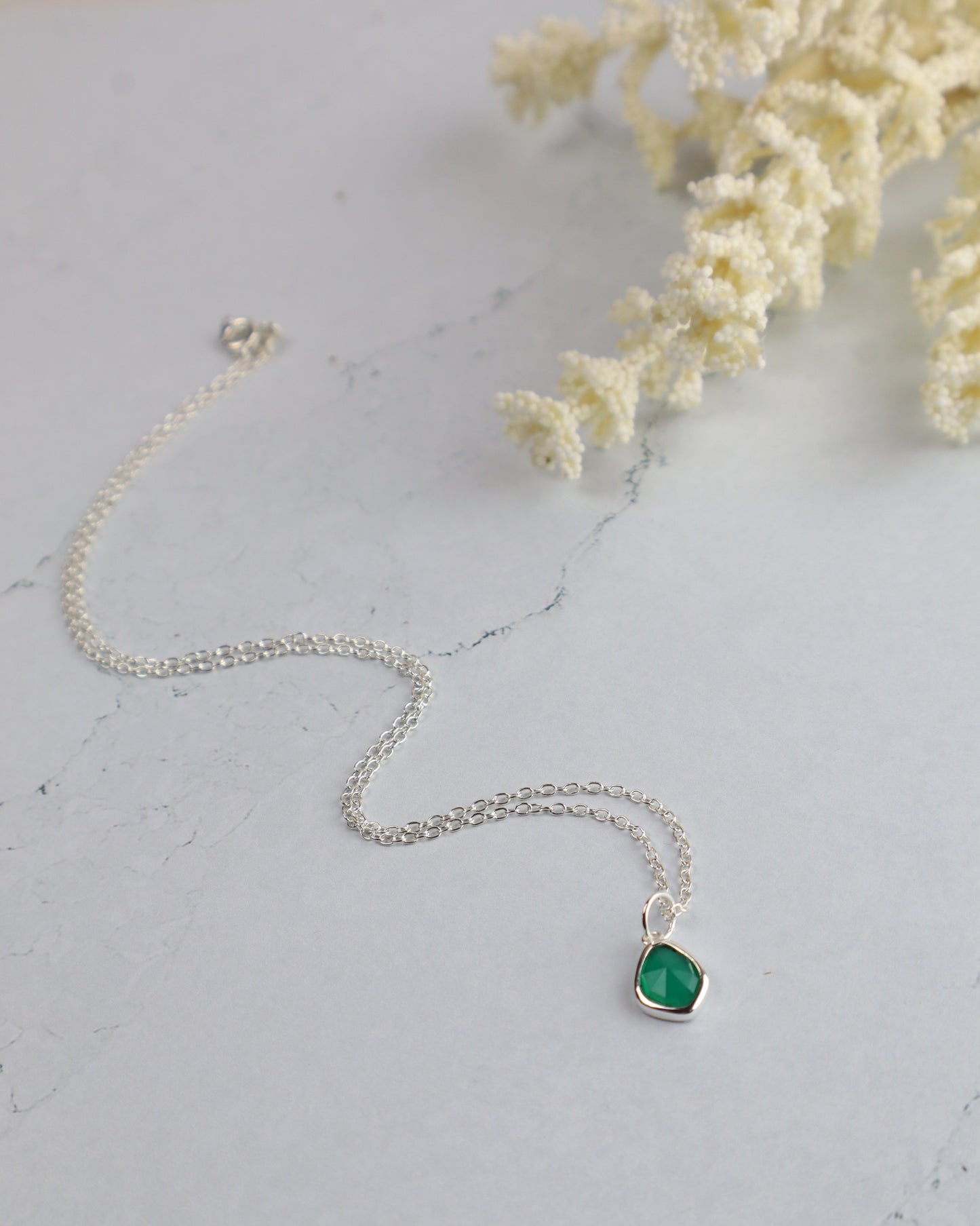 Silver necklace with a green pendant on a light gray surface with white flowers.