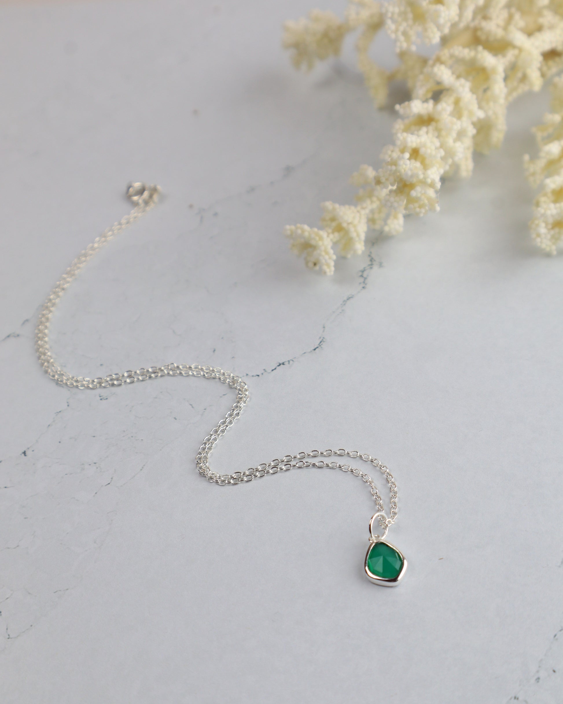 Silver necklace with a green pendant on a light gray surface with white flowers.
