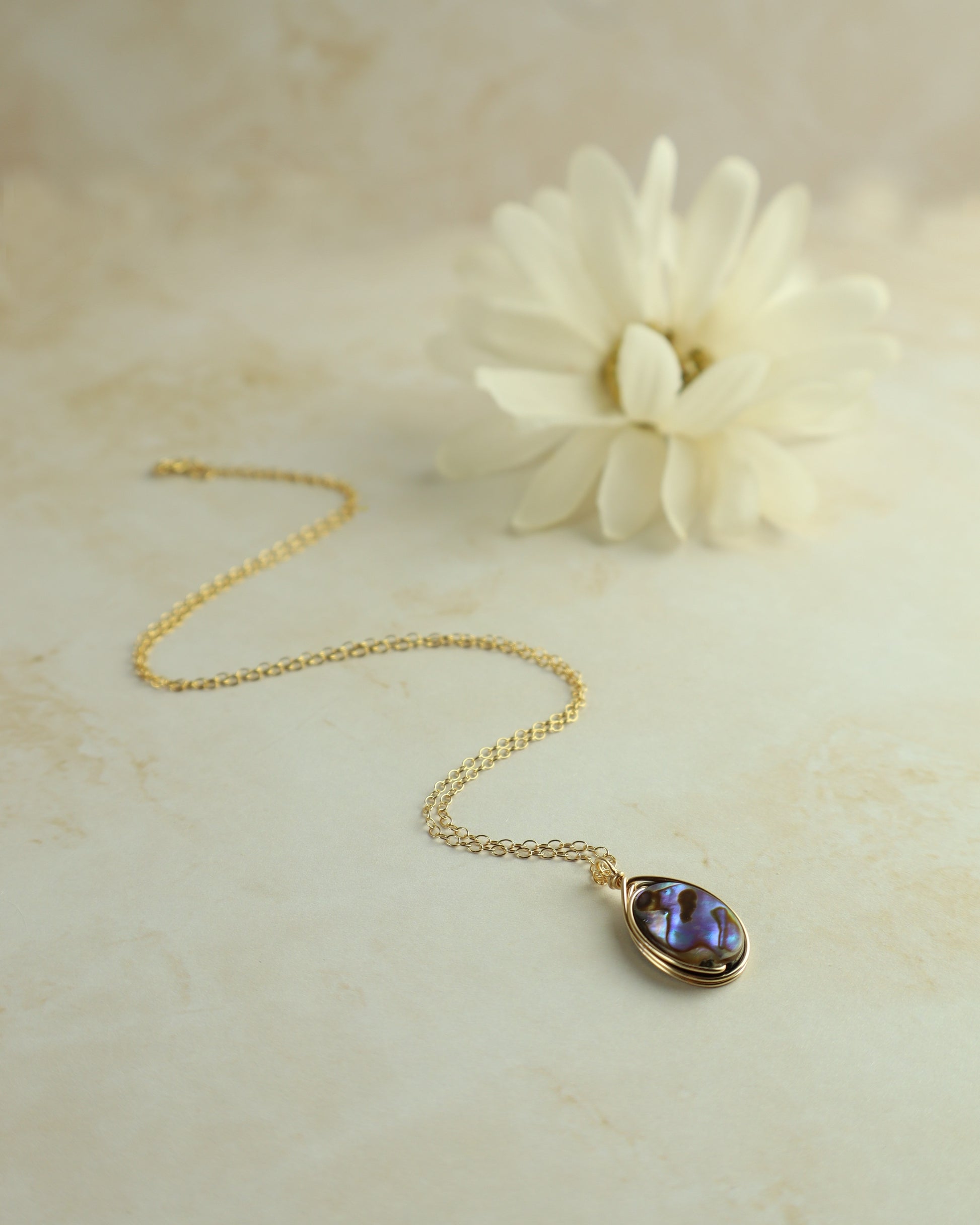 Gold necklace with a purple pendant on a beige surface with a white flower.