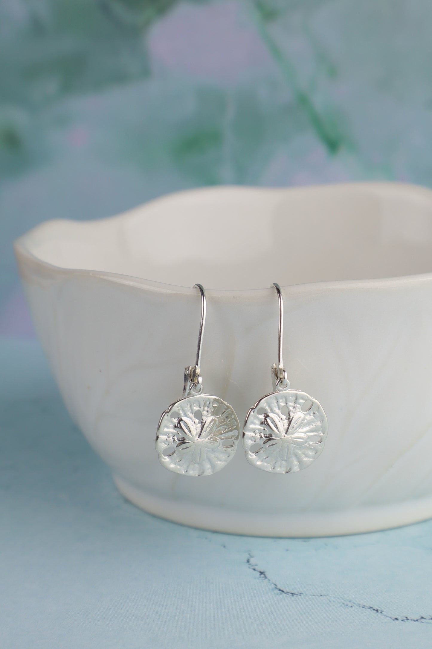 Silver sand dollar earrings on a white dish with a blurred natural background
