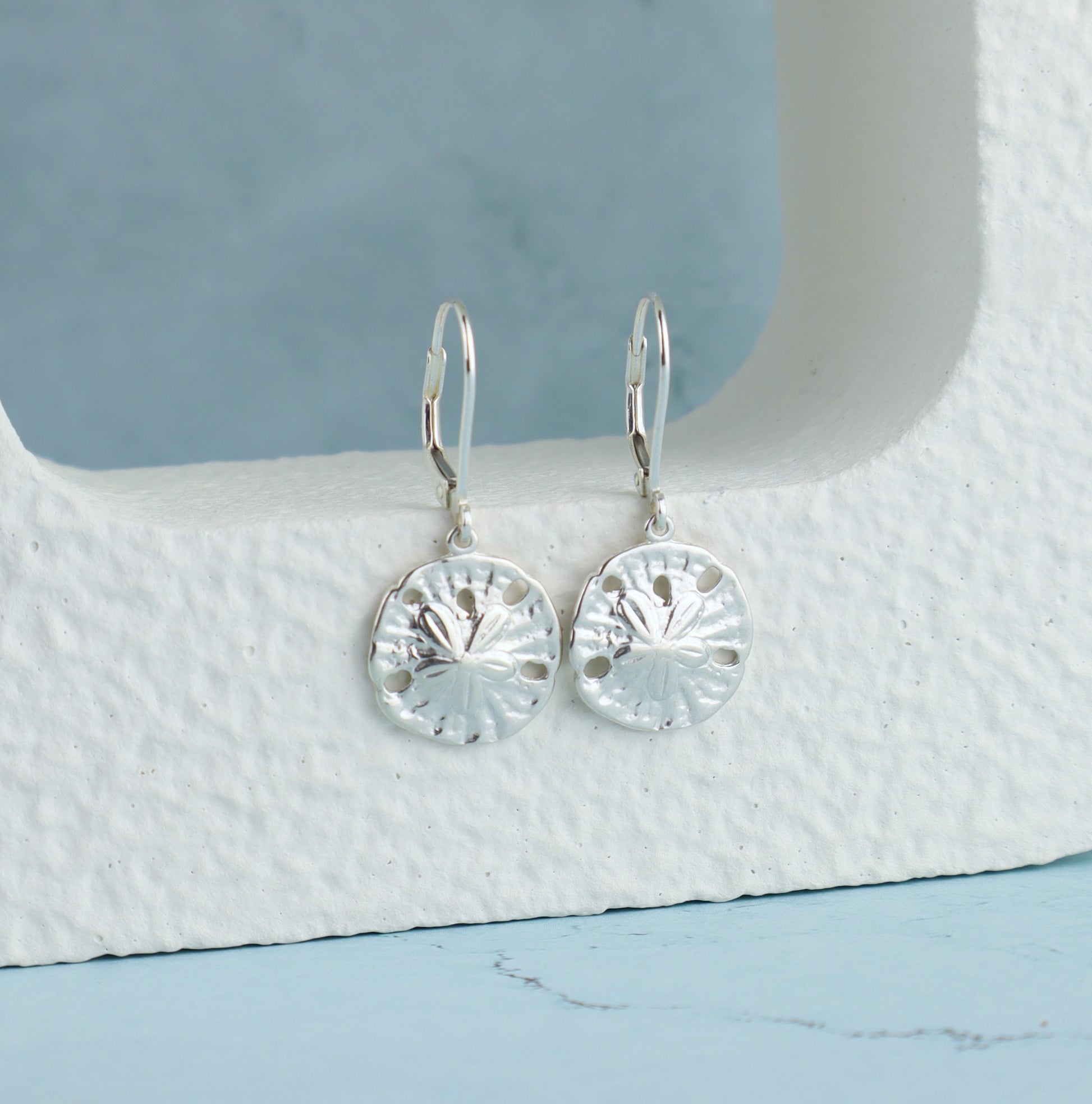 Silver sand dollar earrings on a textured white surface with a blue background
