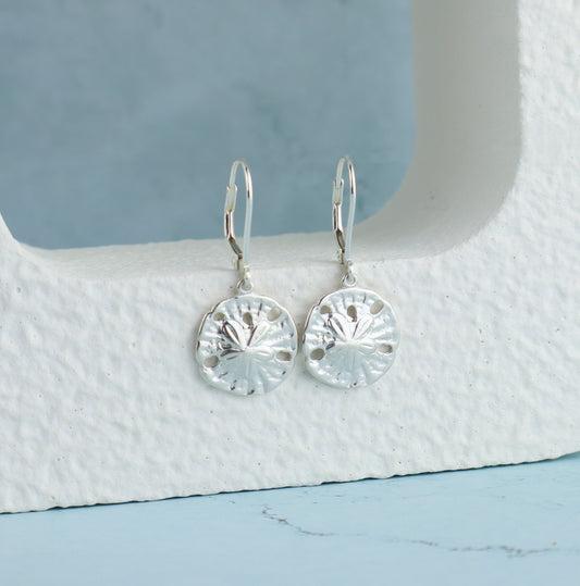 Silver sand dollar earrings on a textured white surface with a blue background