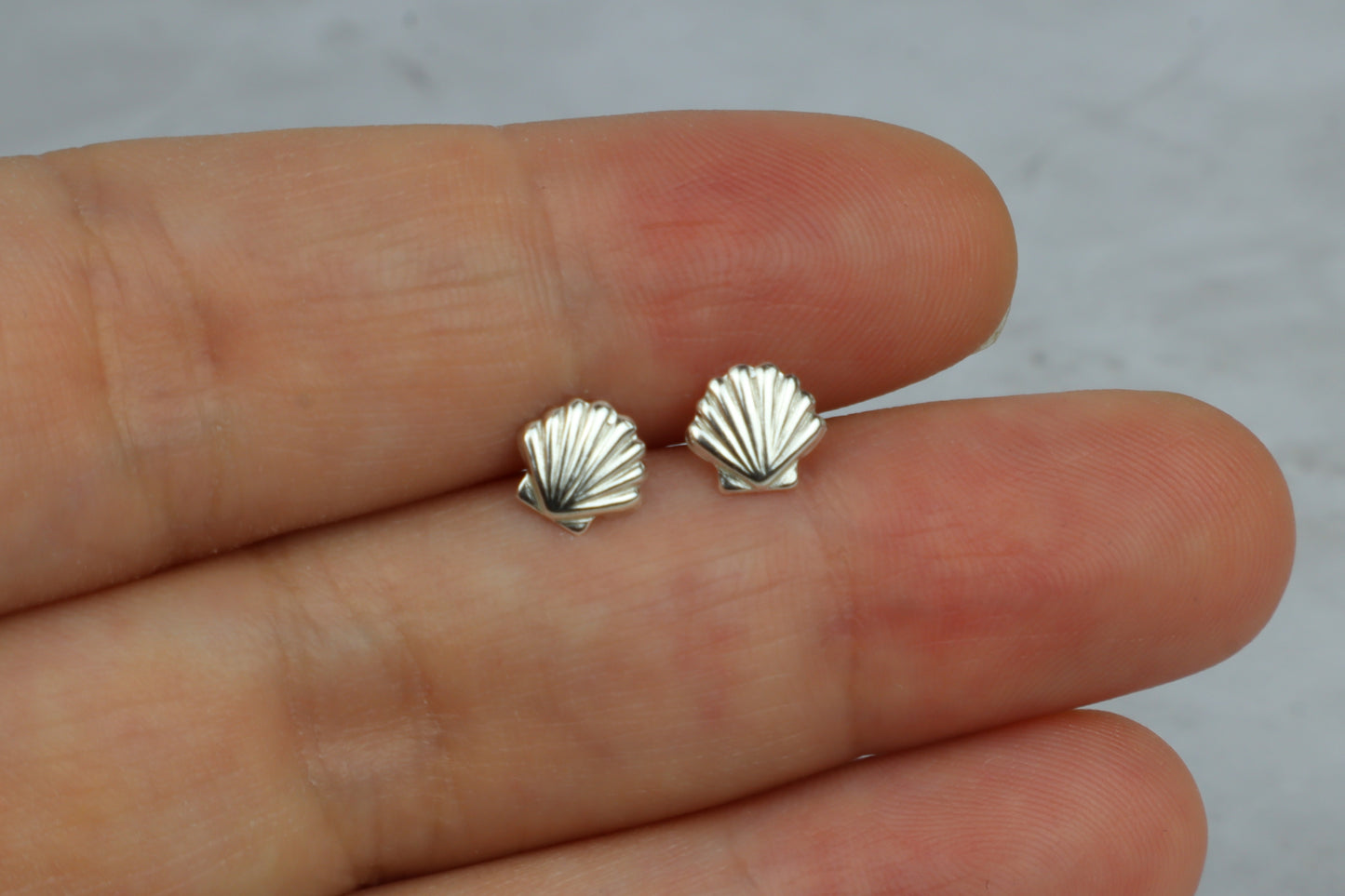 A pair of small silver shell-shaped stud earrings resting on a person's fingers against a light gray background.